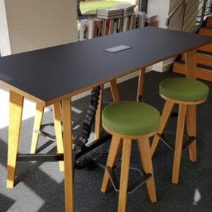 Tall black meeting table with wooden legs and two green upholstered stools in a modern office.