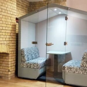 Lismark grey acoustic meeting pod with a house-like roof design, featuring two patterned blue and white upholstered benches, a small table, and clear glass doors, set against an exposed brick wall.